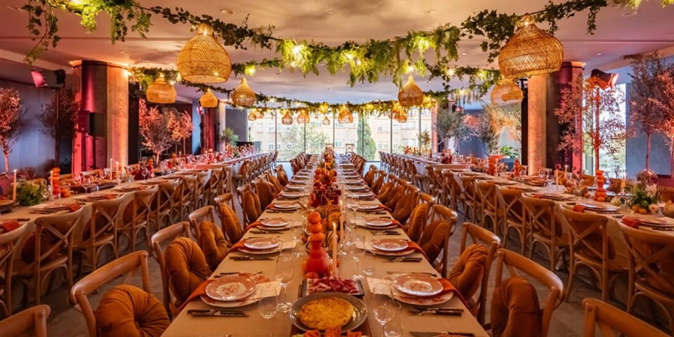 A long banquet table set for a formal dinner, decorated with greenery garlands, hanging lanterns, and warm string lights.