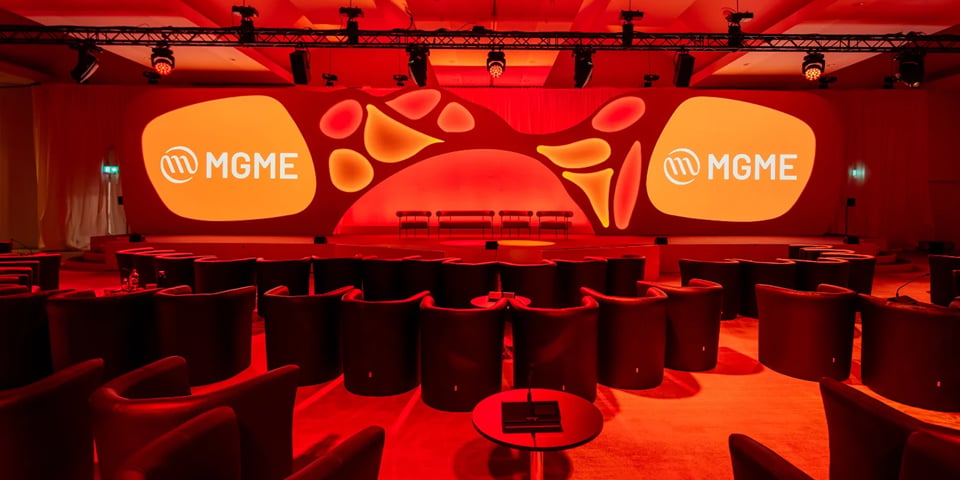 An empty conference stage with curved seating arranged in a circle, lit in red with 'MGME' branding displayed on two large screens.