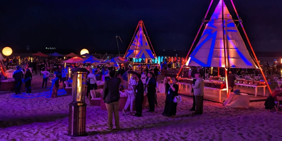 Guests mingle at a nighttime beach event beneath illuminated teepee-style tent structures with blue and purple lighting.