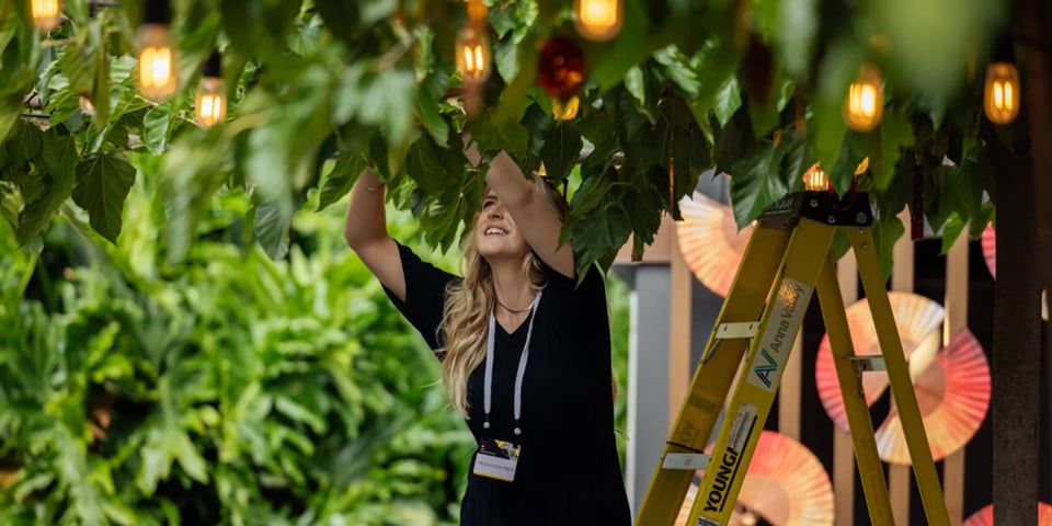 A woman on a ladder adjusts string lights woven through greenery at an outdoor event setup, wearing an event staff lanyard.