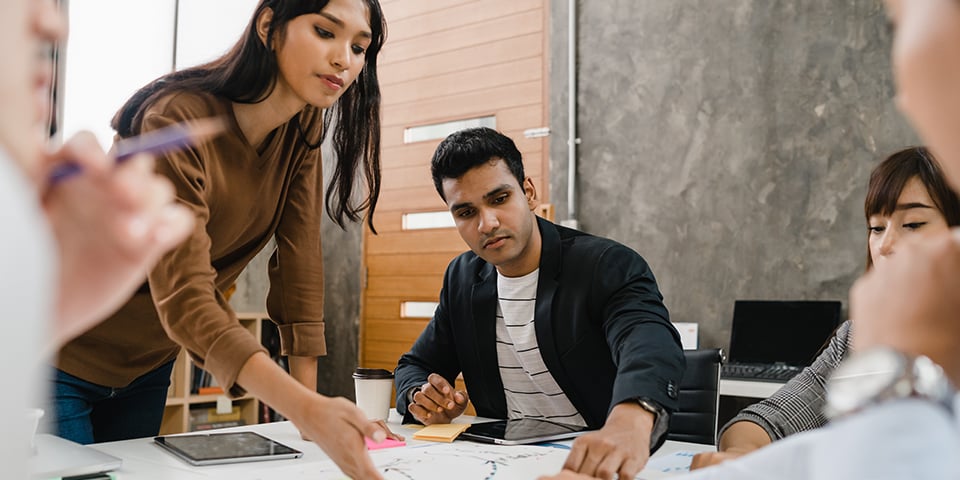 A woman points at a whiteboard sketch while explaining an idea to a seated colleague, with other team members gathered around the table.