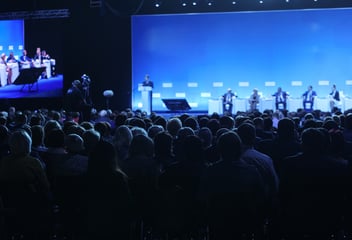 Audience members watch a panel discussion on stage at a large corporate conference, illuminated by blue stage lighting and a big screen.