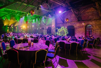 A large corporate event venue with round banquet tables and chairs, set inside an industrial brick hall with dramatic purple and green stage lighting and gold balloons.