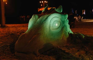A glowing green sand sculpture of a dragon head on a beach at night during an outdoor event.
