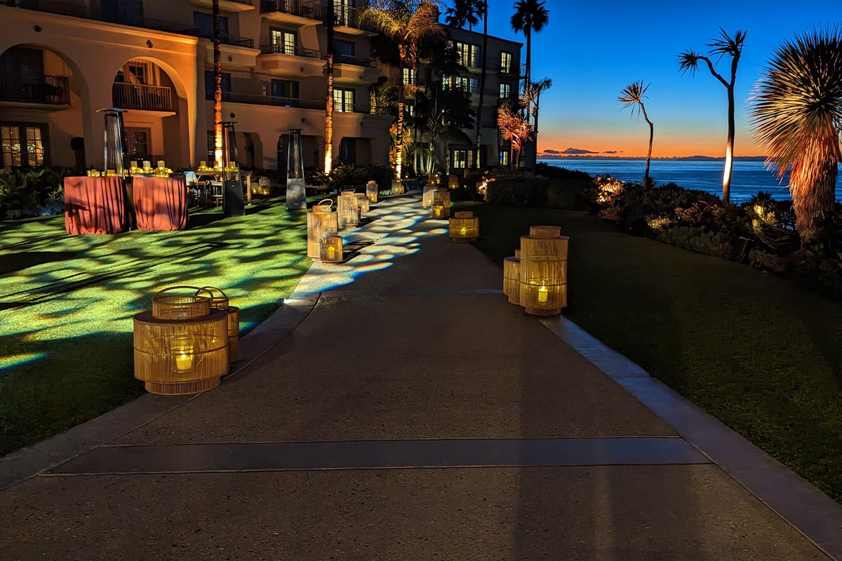 Illuminated oceanfront venue path at sunset with wicker lanterns and green uplighting near palm trees.
