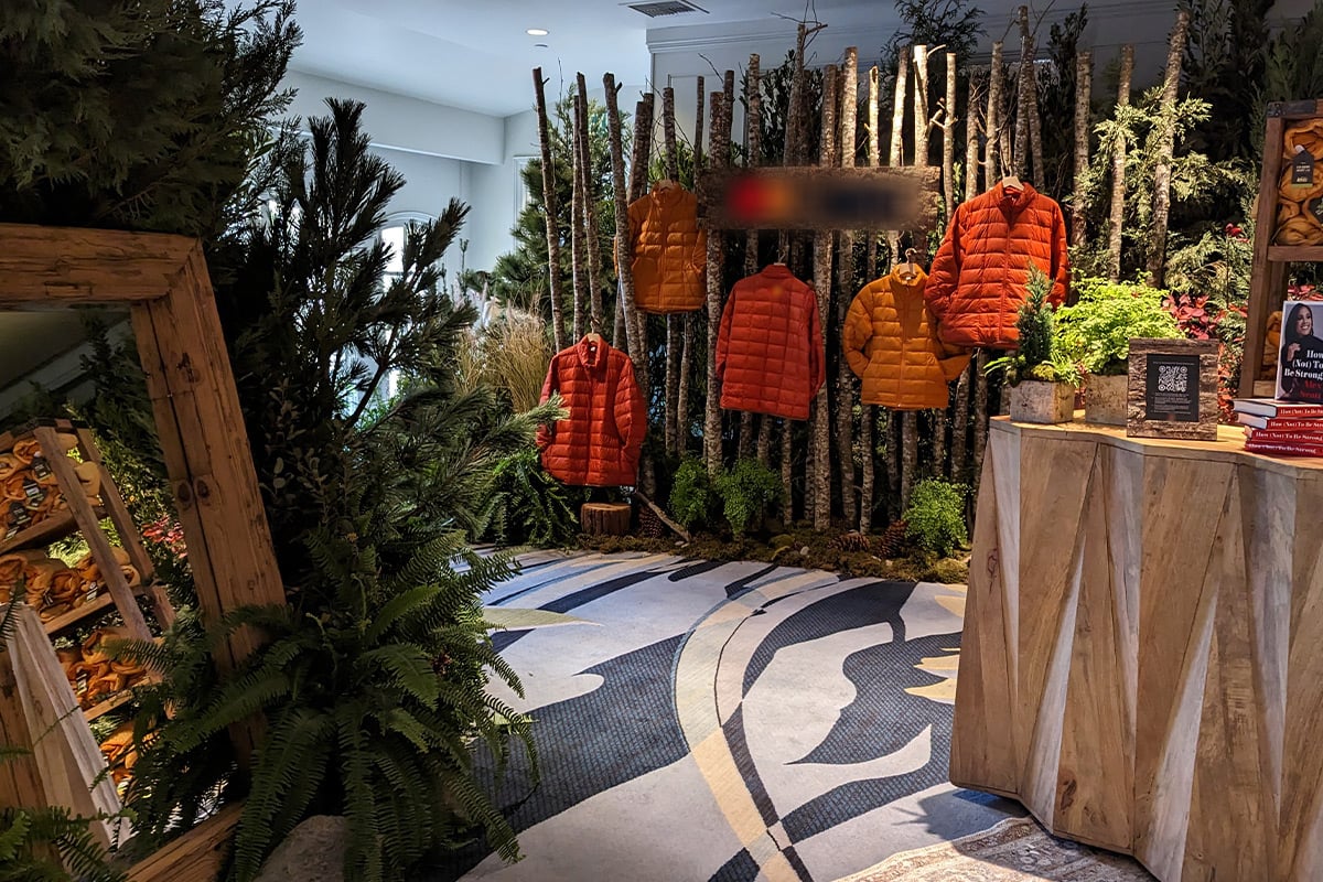 Indoor forest-themed display with orange puffer jackets hanging on birch branches surrounded by ferns and plants.