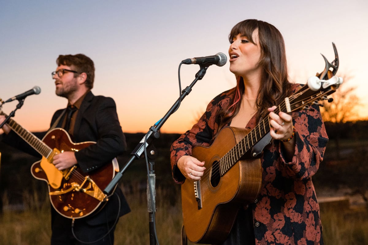 Female singer with acoustic guitar and male guitarist perform at outdoor sunset concert with microphones.