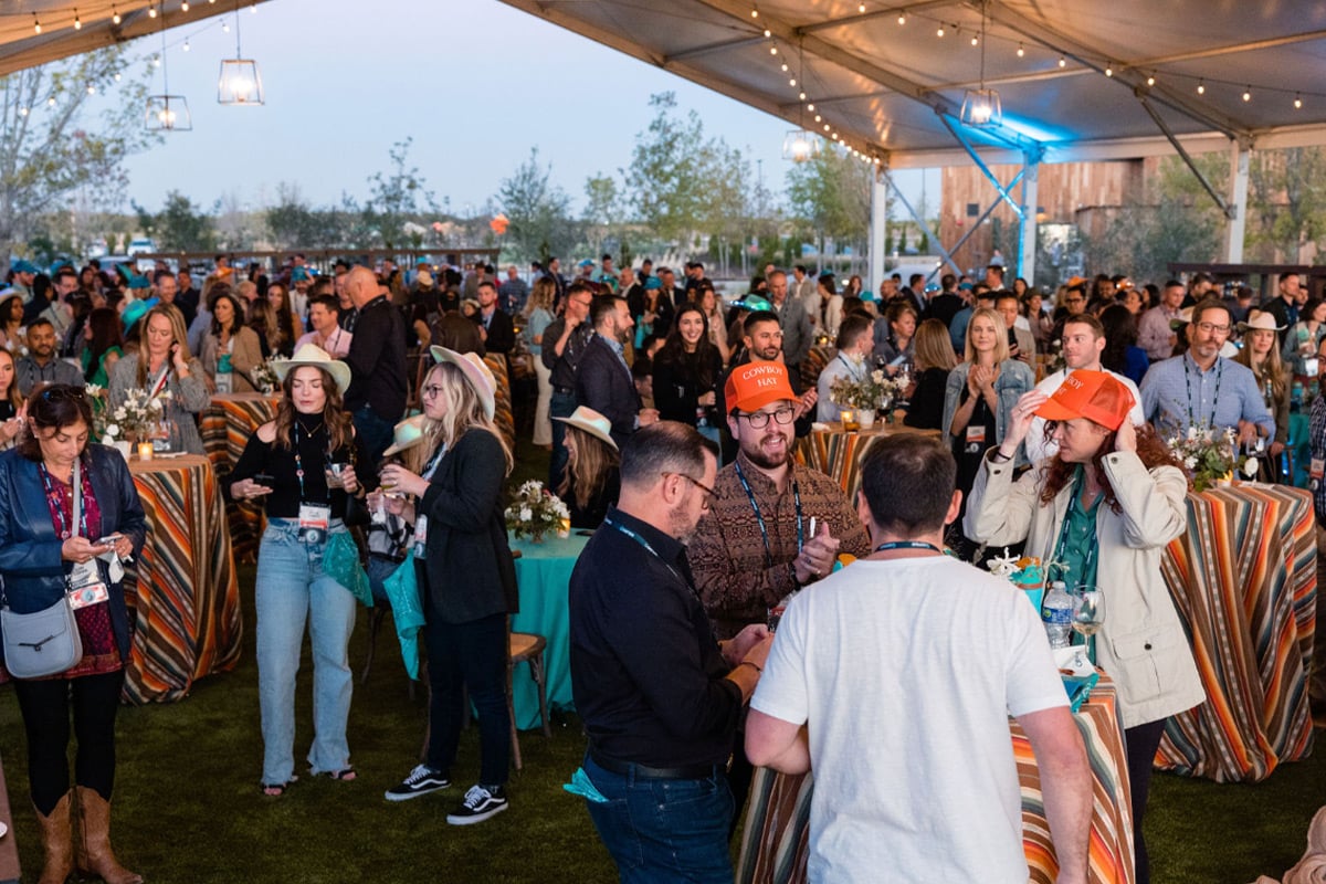 Large crowd mingles at outdoor evening event under tent with string lights and turquoise tablecloths.