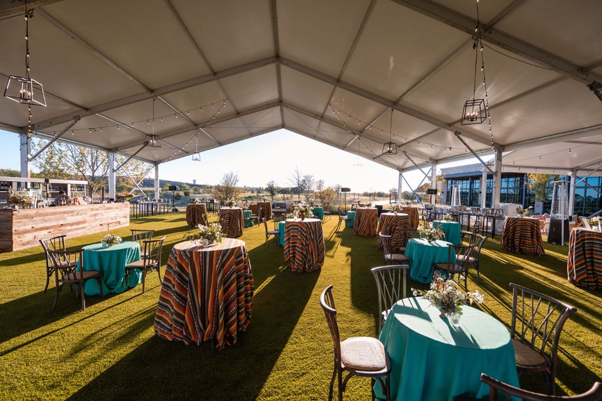 Empty tented outdoor venue with turquoise-clothed cocktail tables and striped runners overlooking green lawn.