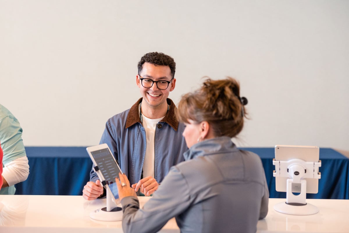 Smiling staff member in glasses assists guest with tablet at modern white check-in desk.