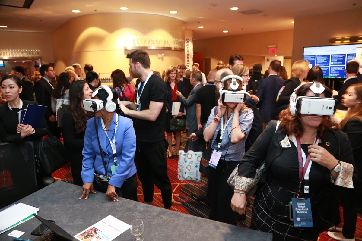 Conference attendees experience virtual reality headsets at crowded exhibition hall with presentation screens visible.