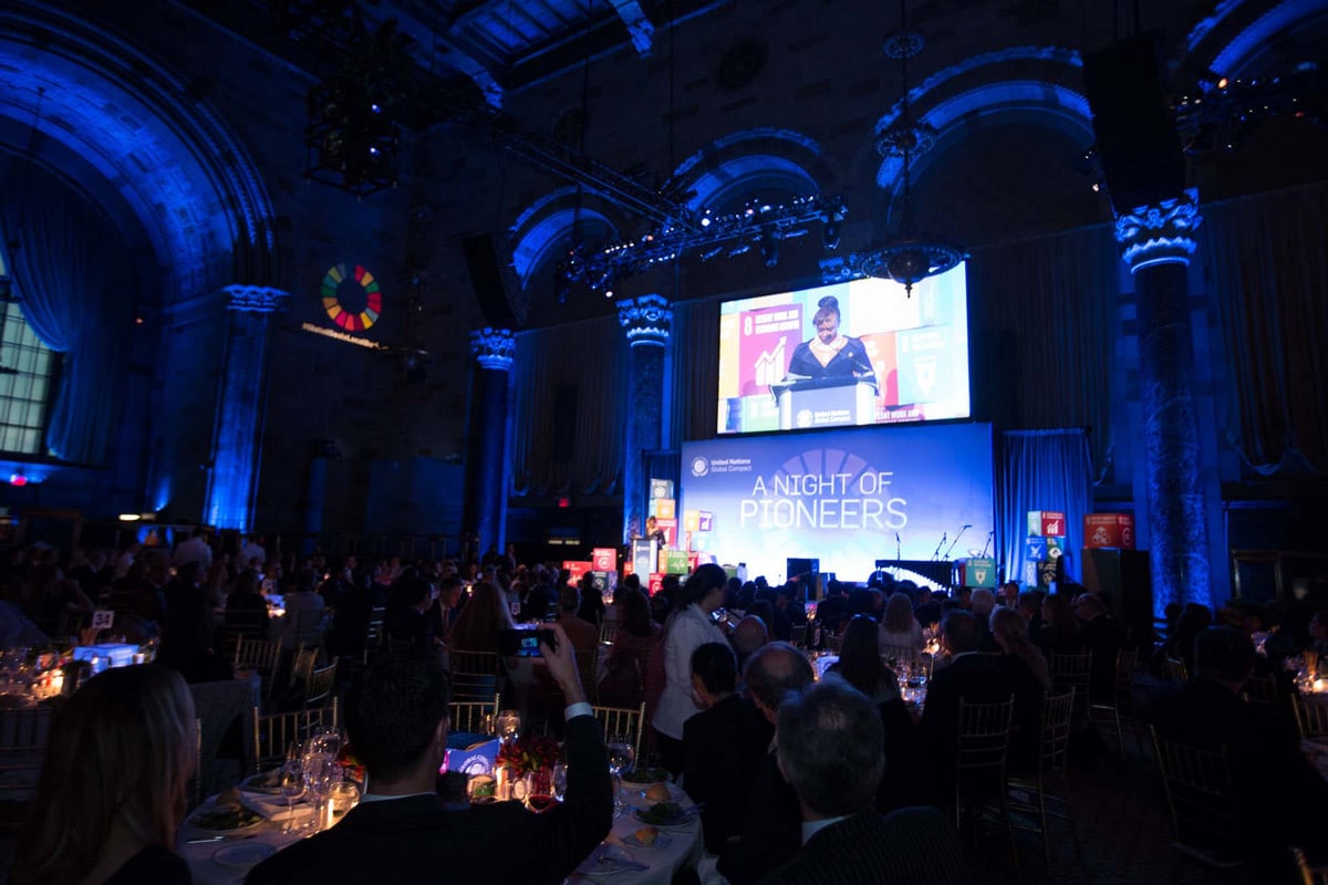 Grand ballroom event with blue uplighting on ornate columns and "A Night of Pioneers" stage presentation.