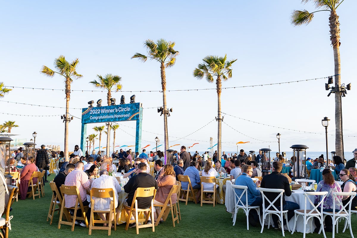 Outdoor dining event with guests at tables under palm trees and string lights near ocean