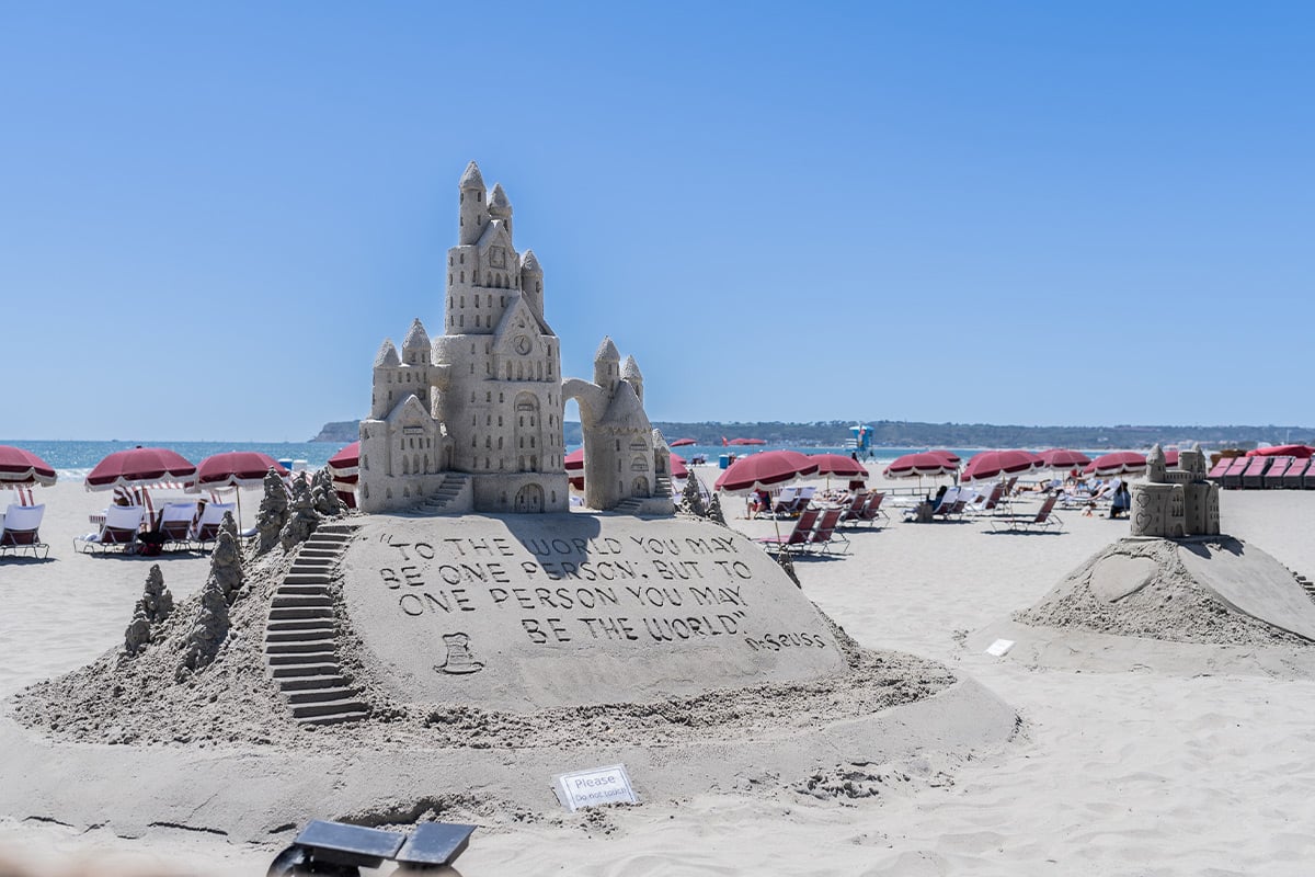 Elaborate sand castle with towers and carved message on sunny beach with pink umbrellas
