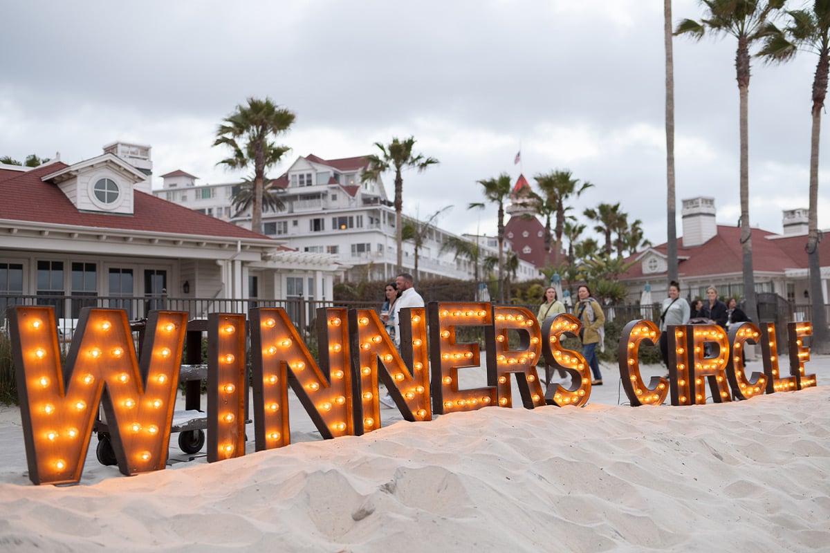 Large marquee letters spelling "WINNERS CIRCLE" illuminated on beach with hotel in background
