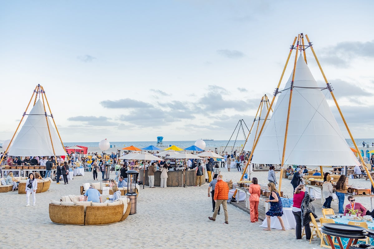Beach event with white teepee tents, colorful umbrellas, and guests mingling on sand near ocean